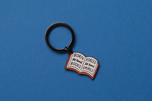 An enamel keychain of an open book that says "Born to read books, Forced to read emails" over a blue background.