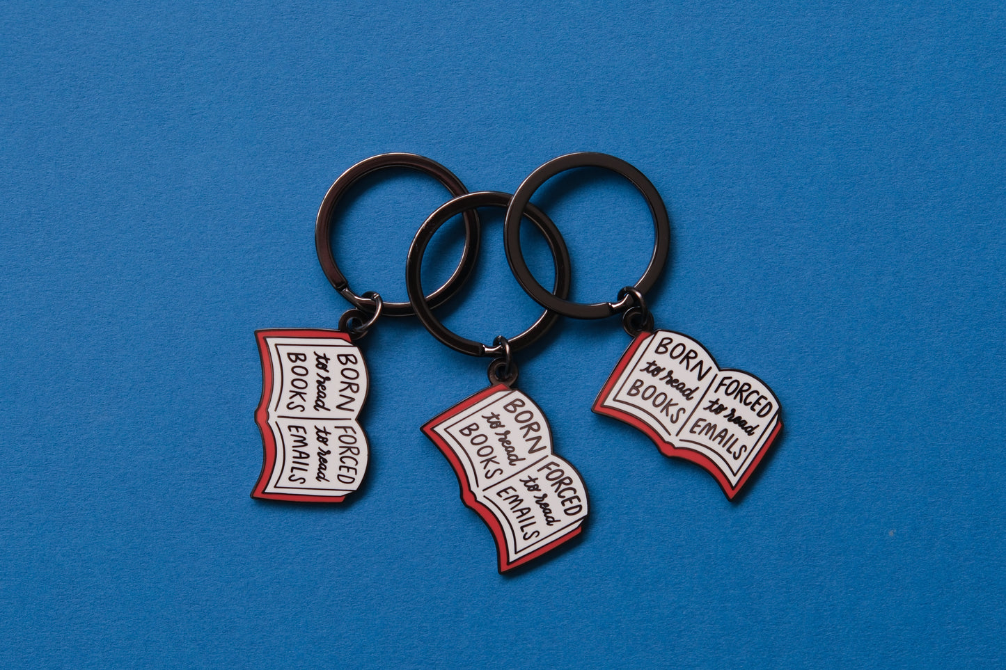 3 enamel keychains of open books that say "Born to read books, Forced to read emails" over a blue background.