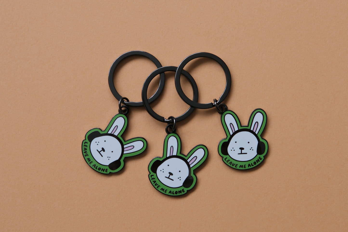 3 enamel keychains of rabbits wearing headphones that say "Leave me alone" over an orange background.