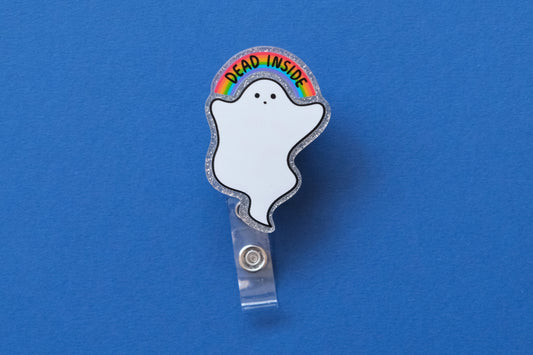 Photo of a silver glitter acrylic badge reel shaped like a ghost with a rainbow that says "Dead Inside" over a dark blue background.
