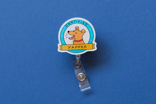 Photo of an acrylic badge reel shaped like a dog badge that says "Certified Yapper" over a blue background.
