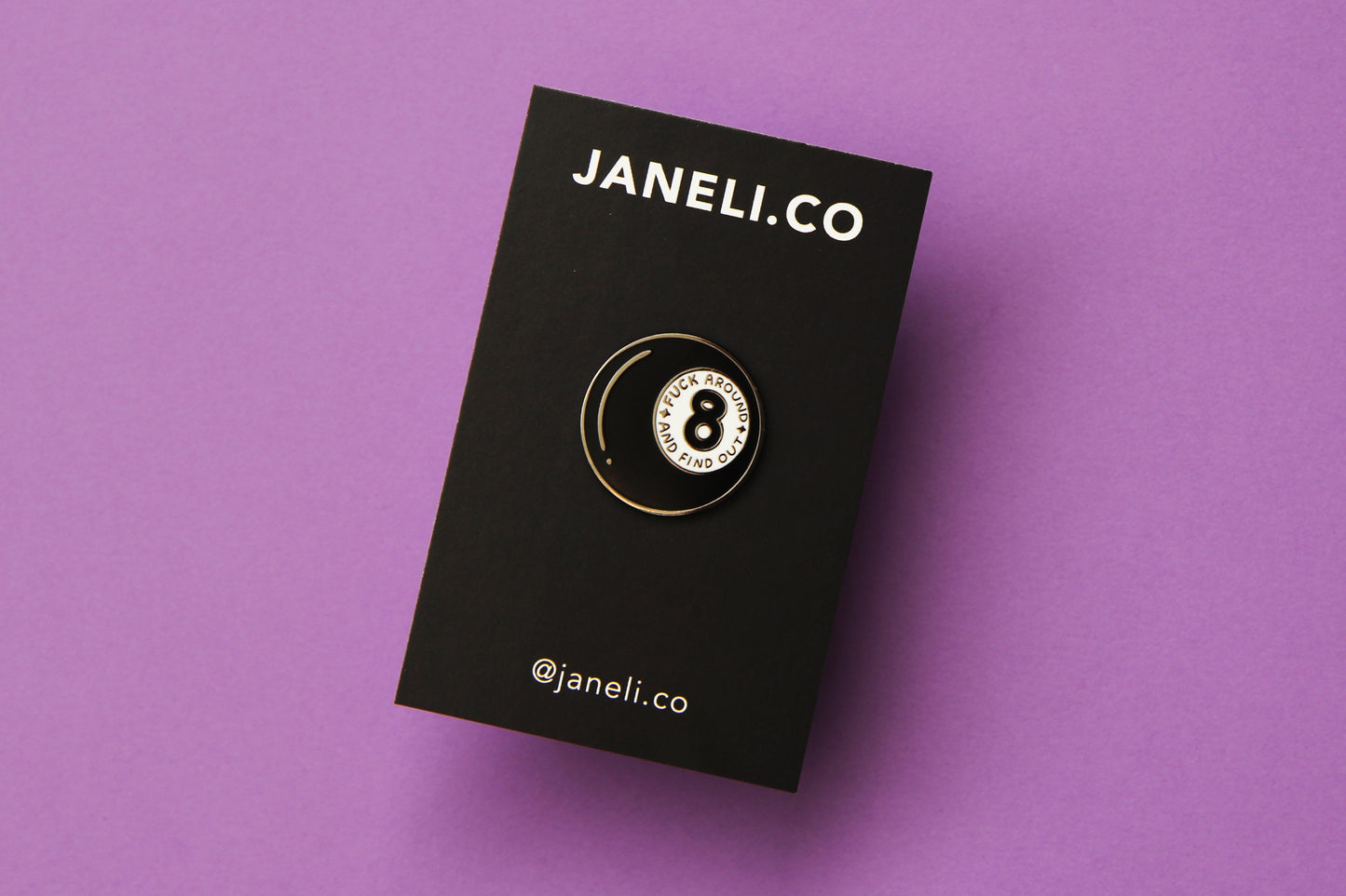 An enamel pin showing a magic 8 ball that says "Fuck around and find out" on a black JaneLi.Co backing card over a purple background.