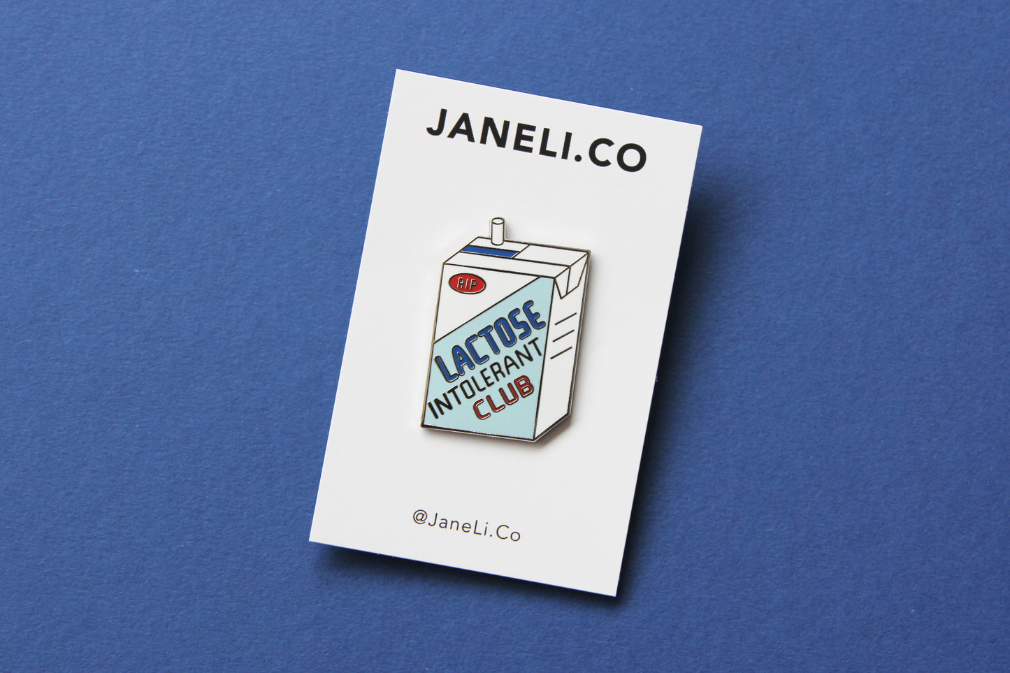 An enamel pin showing a soymilk carton that says" Lactose Intolerant Club" on a white JaneLi.Co backing card over a blue background.