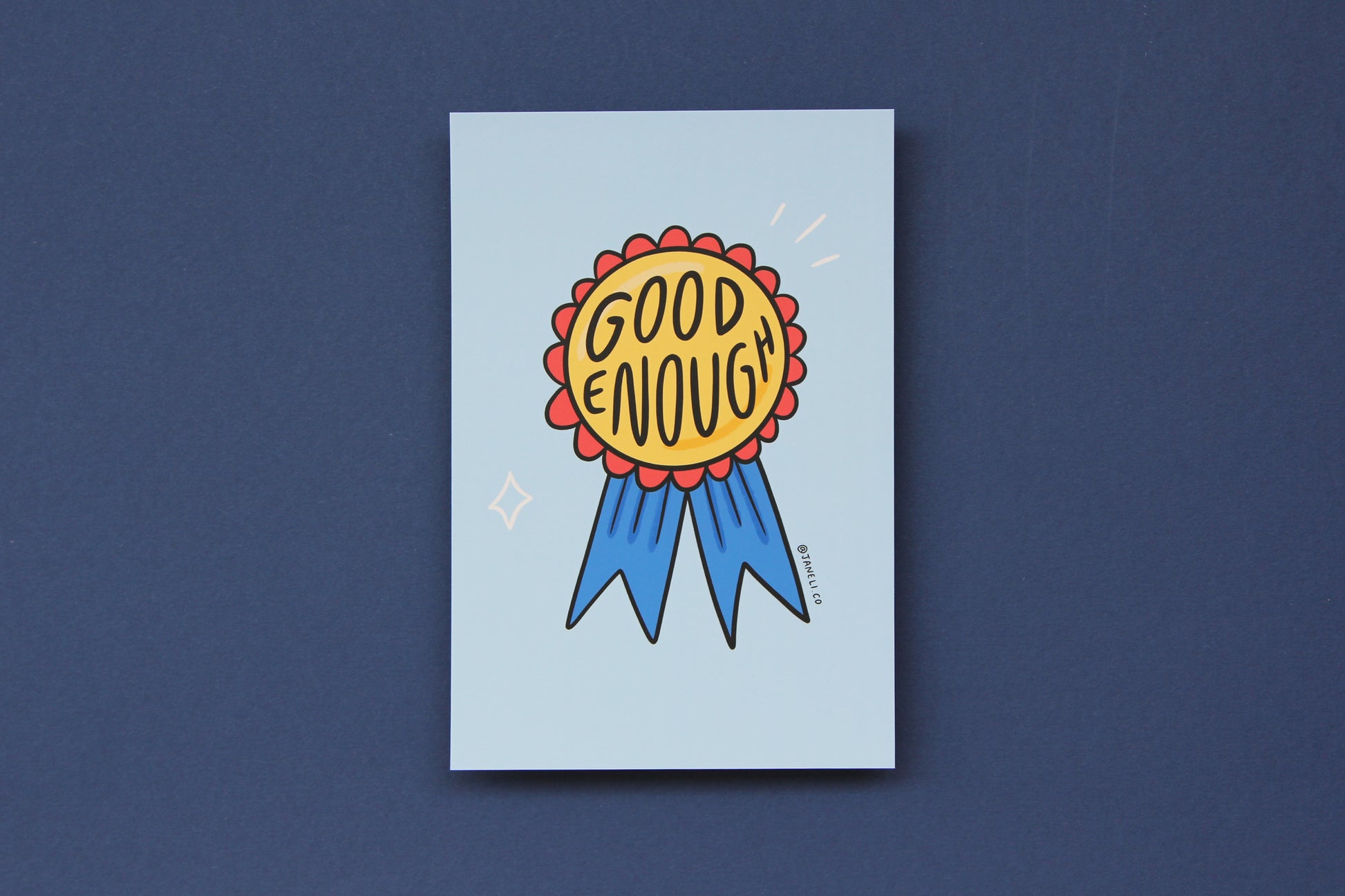 A JaneLi.Co mini print/postcard of an award ribbon that says "Good Enough" over a blue background.
