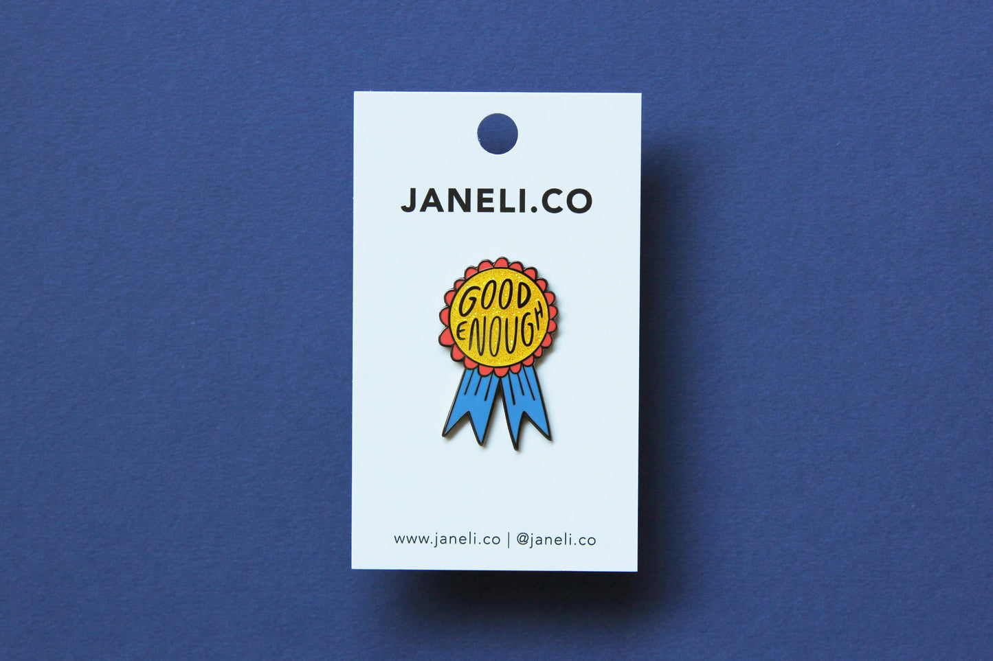 An enamel pin showing a glittery red and blue award ribbon that says "Good Enough" on a white JaneLi.Co backing card over a blue background.