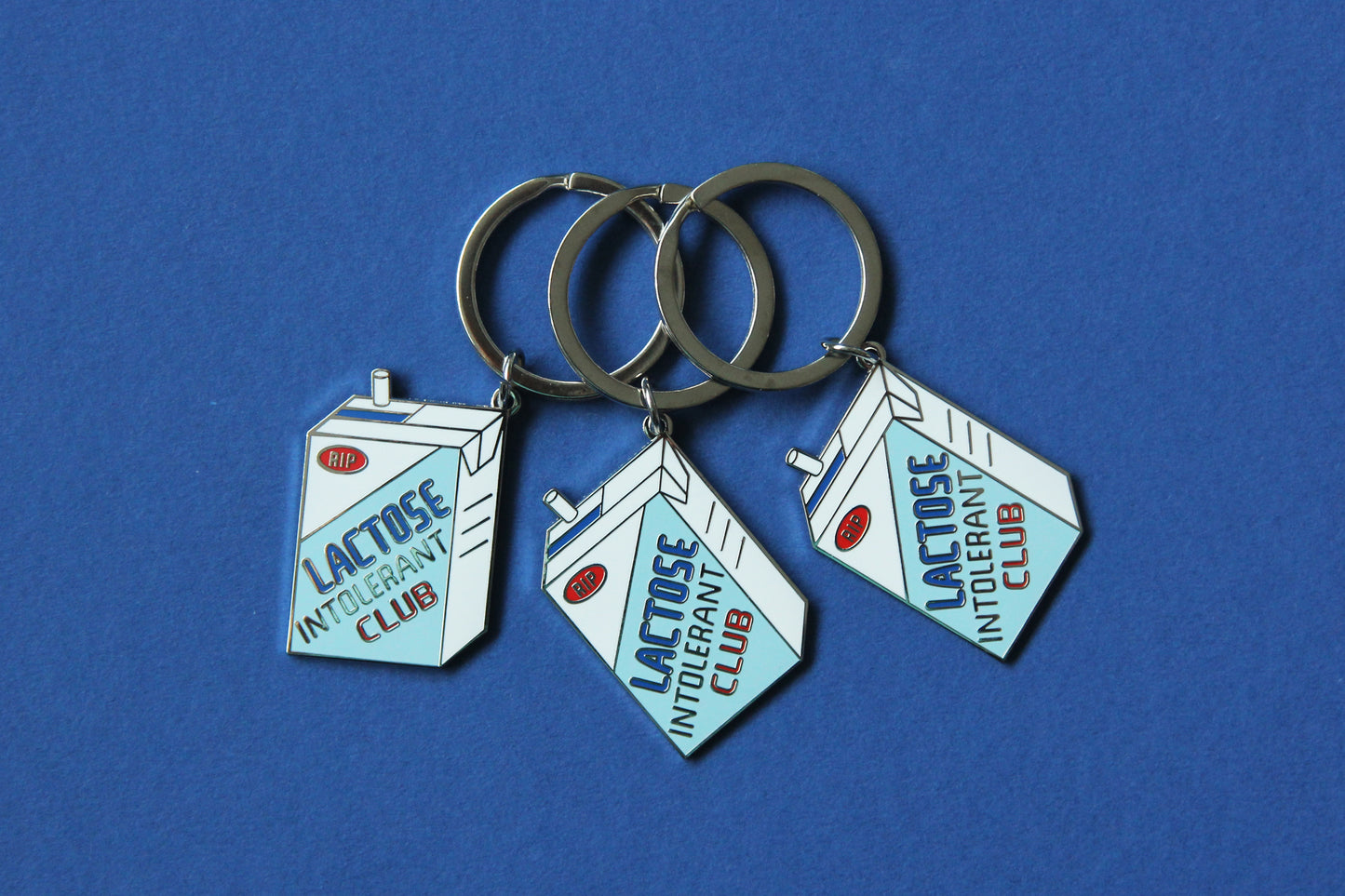 3 enamel keychains showing a soymilk carton that says" Lactose Intolerant Club" over a blue background.