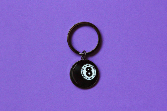 An enamel keychain showing a magic 8 ball that says "Fuck around and find out" over a purple background.