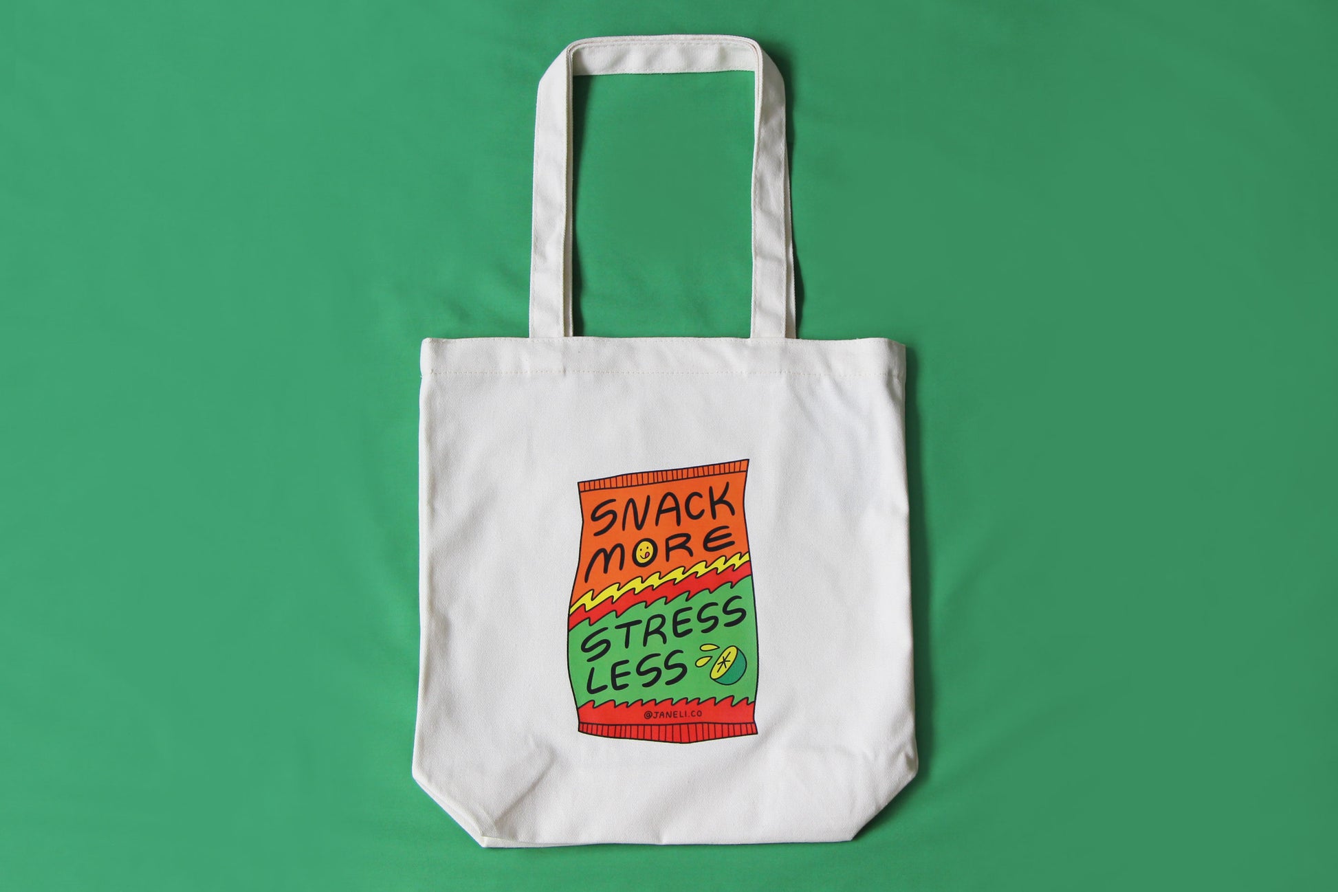 A photo of a natural canvas tote with a chip bag that says "Snack More Stress Less" on it over a green background.