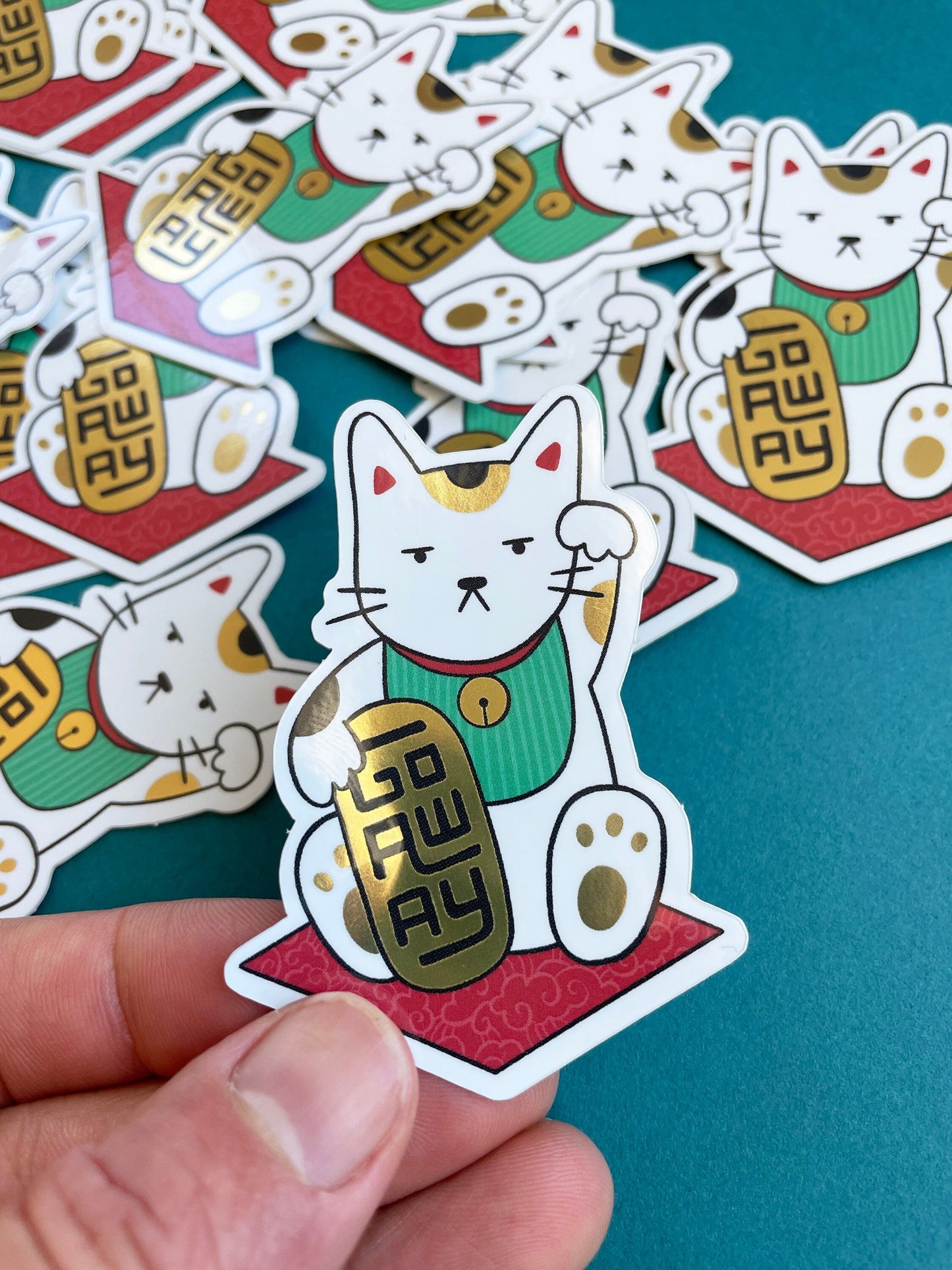 A hand holding a JaneLi.Co sticker that shows a Maneki Neko cat holding a metallic gold bar that says "Go Away" over a pile of Maneki Neko stickers on a teal background.