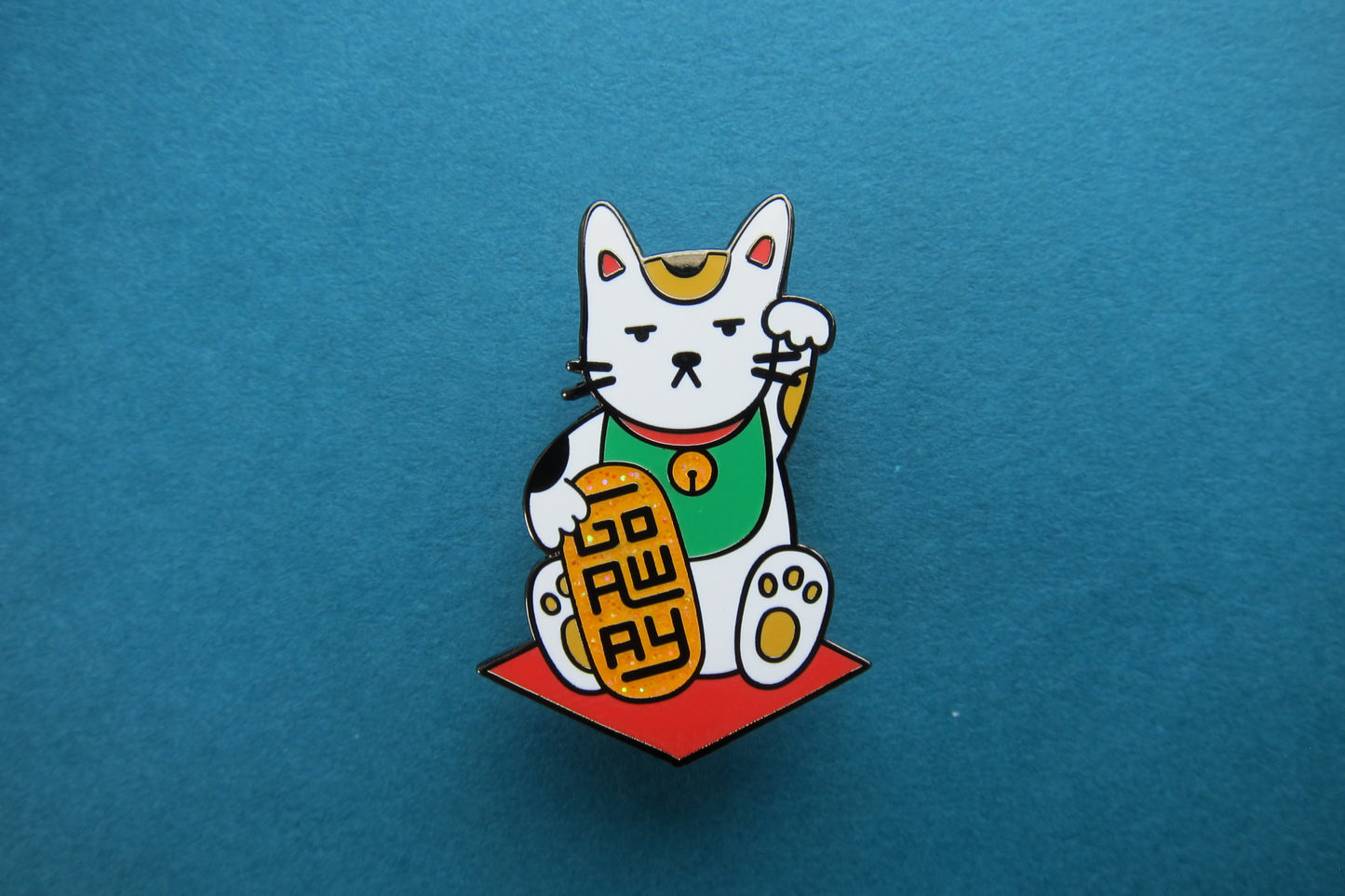 An enamel pin showing maneki neko cat holding a glittery gold bar that says "Go Away" over a teal background.