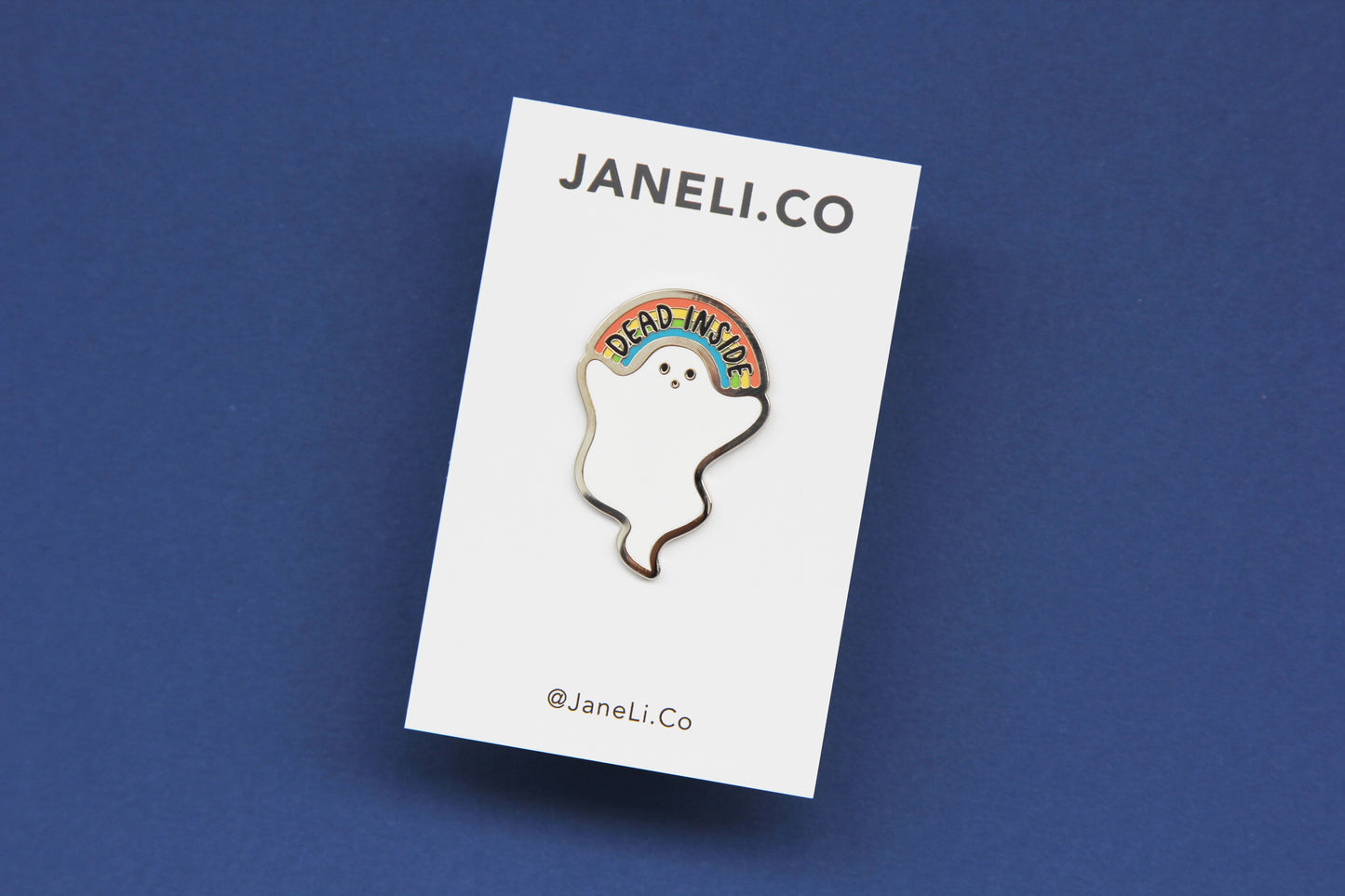 An enamel pin showing a little white ghost holding a rainbow that says "Dead Inside" on a white JaneLi.Co backing card over a navy background.