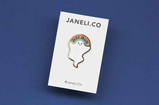 An enamel pin showing a little white ghost holding a rainbow that says "Dead Inside" on a white JaneLi.Co backing card over a navy background.