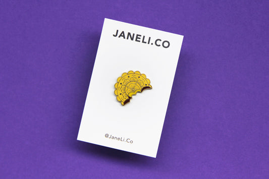 An enamel pin showing a mooncake with a bite out of it on a white JaneLi.Co backing card over a purple background.