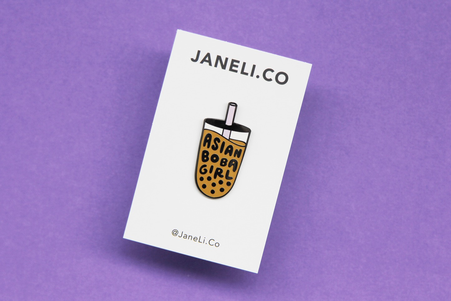 An enamel pin showing a cup of boba that says "Asian Boba Girl" with a purple straw on a white JaneLi.Co backing card over a purple background.