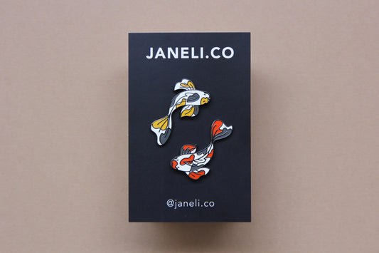 A set of 2 koi fish enamel pins circling on a black JaneLi.Co backing card over a tan background. One koi fish is gold and grey, and the other is scarlet and grey.