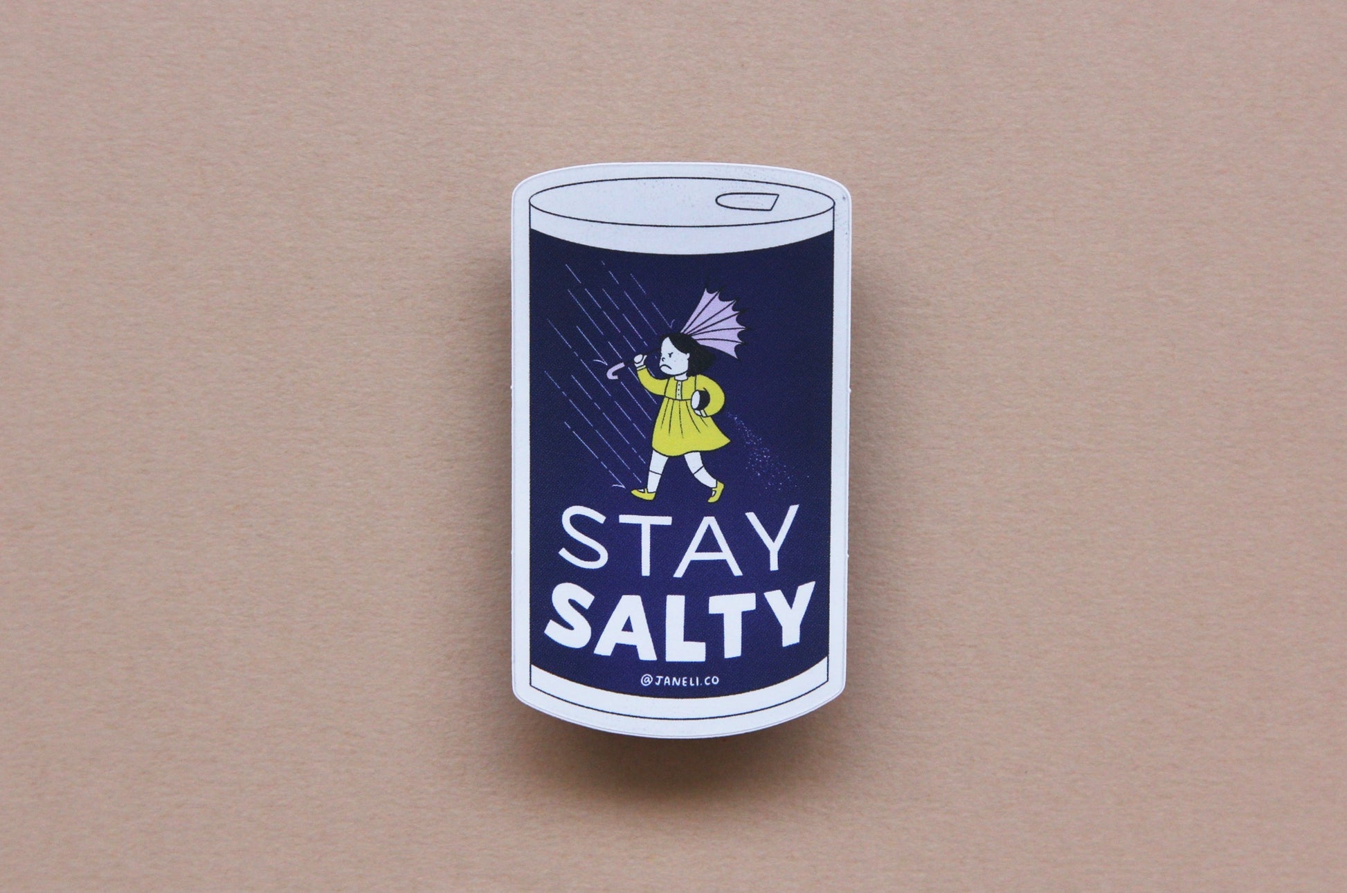 A JaneLi.Co sticker that says "Stay Salty" in the shape of a salt can with an angry salt girl holding an umbrella blow backwards from wind and rain over a navy blue background.