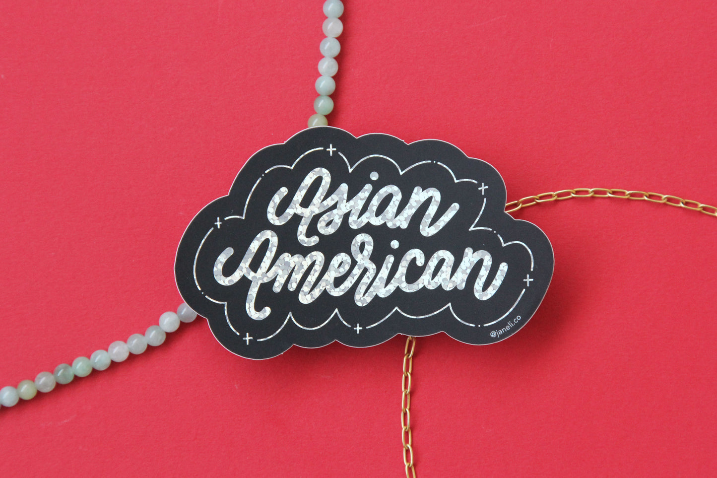 A black and metallic glitter JaneLi.Co sticker that says "Asian American" in cursive lettering over a red background with a string of green jade and a thin gold chain.