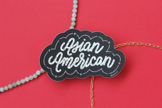 A black and metallic glitter JaneLi.Co sticker that says "Asian American" in cursive lettering over a red background with a string of green jade and a thin gold chain.