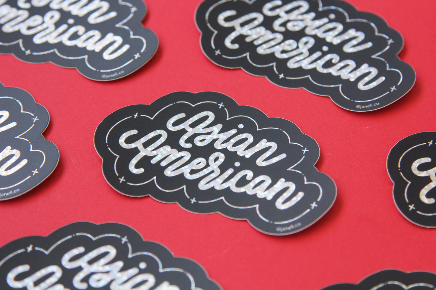 A grid of black and metallic glitter JaneLi.Co stickers that say "Asian American" in cursive lettering over a red background.