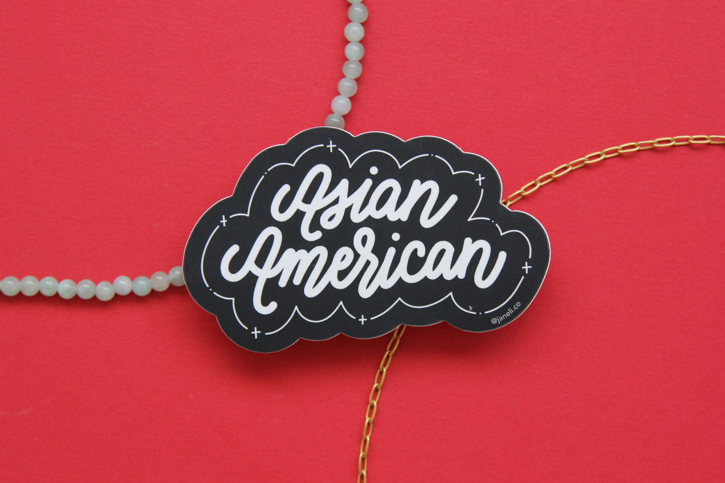 A black and white JaneLi.Co sticker that says "Asian American" in cursive lettering over a red background with a string of green jade and a thin gold chain.