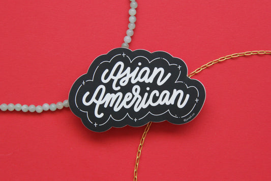 A black and white JaneLi.Co sticker that says "Asian American" in cursive lettering over a red background with a string of green jade and a thin gold chain.