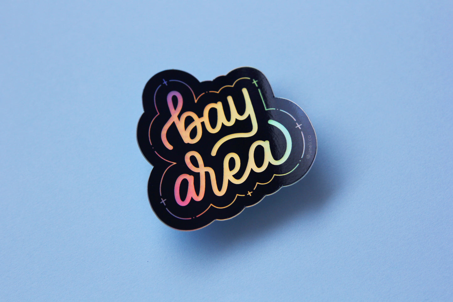 A holographic JaneLi.Co sticker that says "Bay Area" in cursive lettering over a sky blue background.