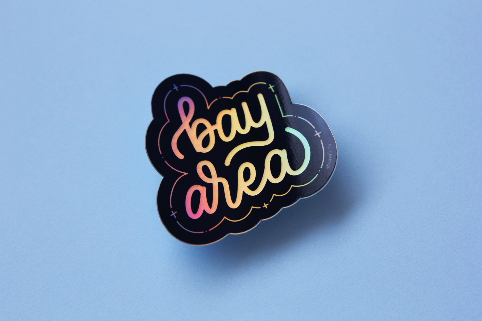 A holographic JaneLi.Co sticker that says "Bay Area" in cursive lettering over a sky blue background.