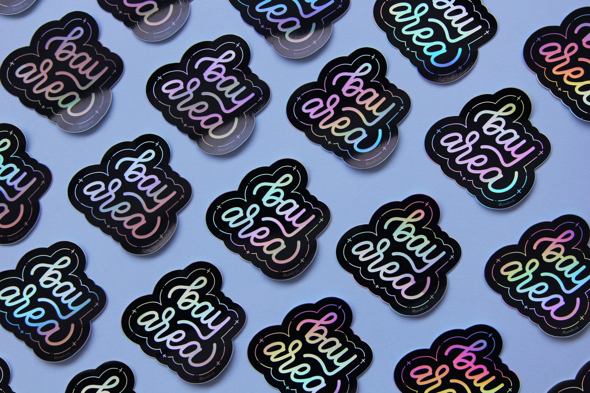 A grid of holographic JaneLi.Co stickers that say "Bay Area" in cursive lettering over a sky blue background.