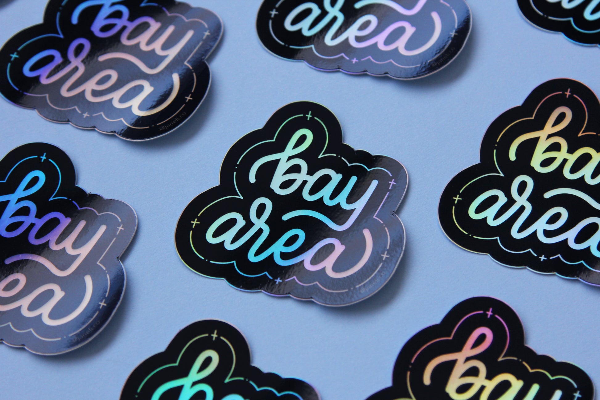 A grid of holographic JaneLi.Co stickers that say "Bay Area" in cursive lettering over a sky blue background. 