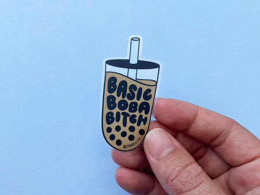 A hand holding a clear JaneLi.Co sticker that says "Basic Boba Bitch" in the shape of a cup of boba over a sky blue background.
