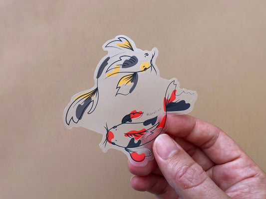 A hand holding a clear JaneLi.Co sticker of a gold and grey koi fish and a scarlet and black koi fish swimming in a circle over a tan background. 