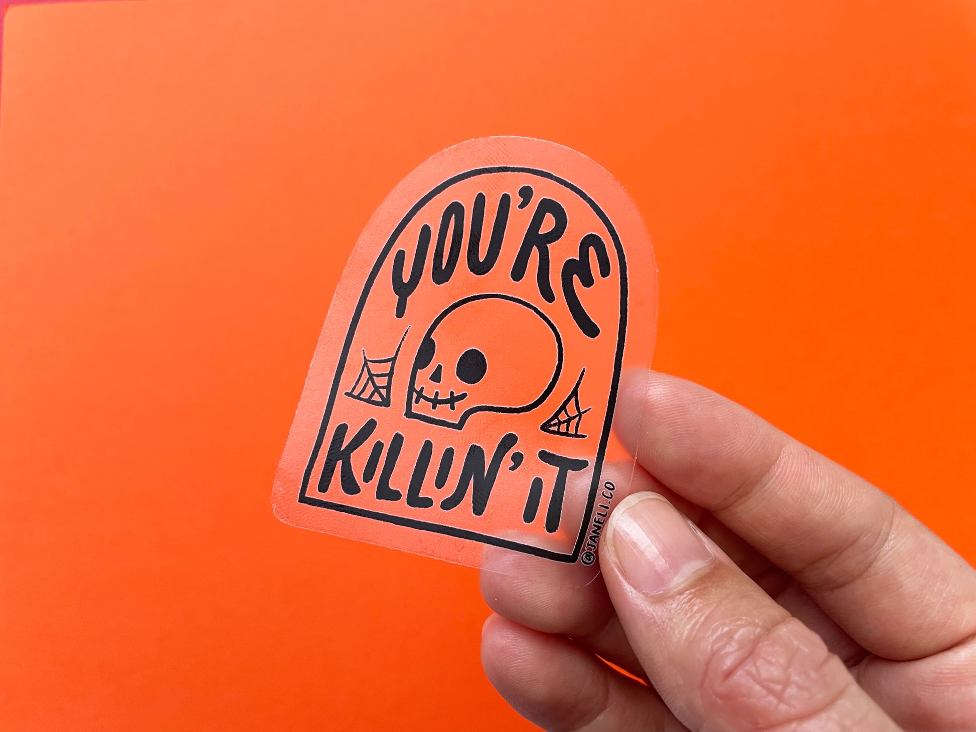 A hand holding a clear JaneLi.Co sticker that says "You're Killin' It" in the shape of a tombstone with a skull in it over an orange background.