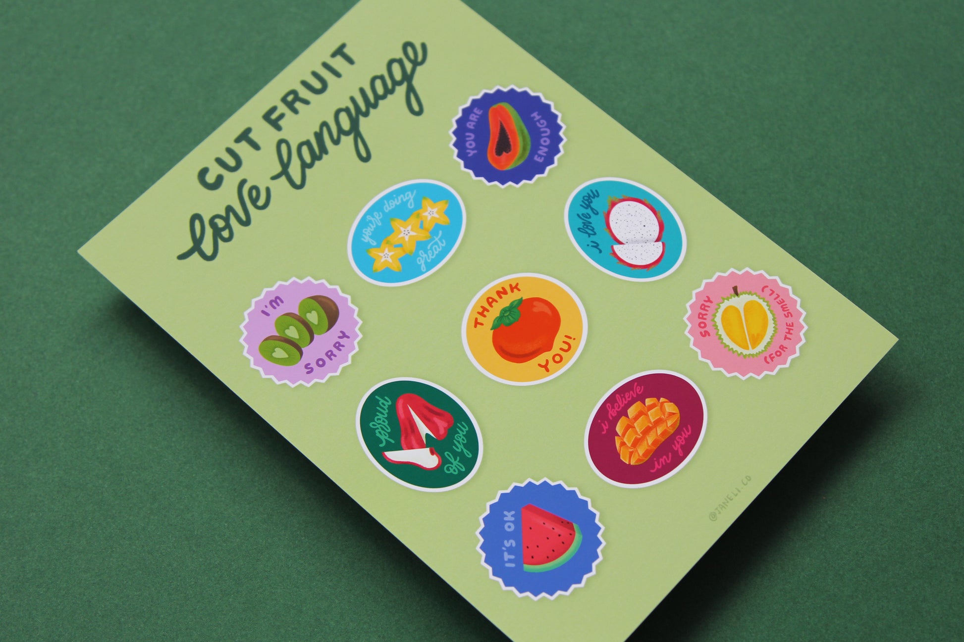 A close up of a JaneLi.Co print that says "Cut Fruit Love Language" with 9 different fruit stickers with different messages like "I love you", "I'm sorry", and "I'm proud of you" over a green background.