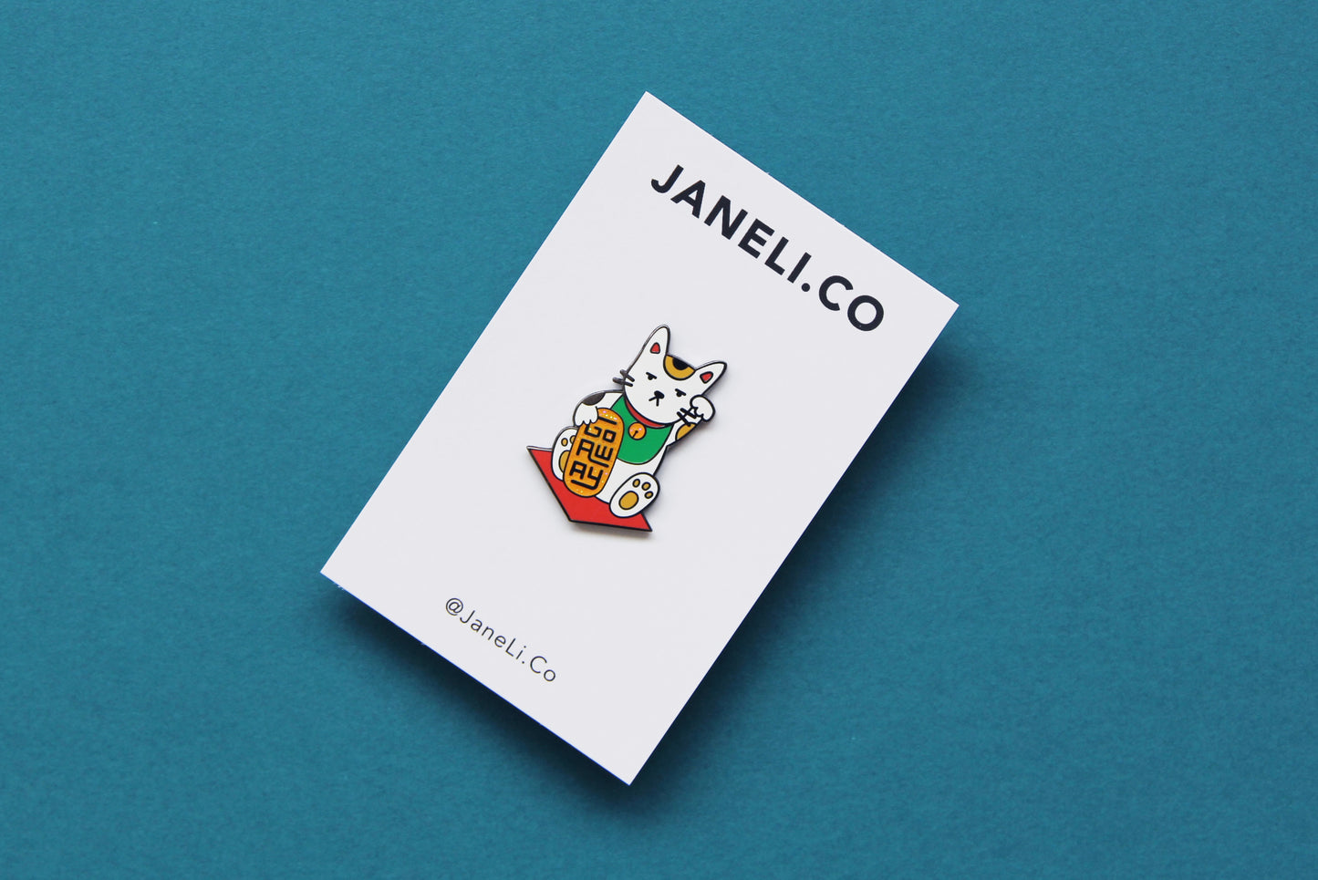 An enamel pin showing maneki neko cat holding a glittery gold bar that says "Go Away" on a white JaneLi.Co backing card over a teal blackground.