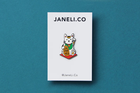 An enamel pin showing maneki neko cat holding a glittery gold bar that says "Go Away" on a white JaneLi.Co backing card over a teal blackground.