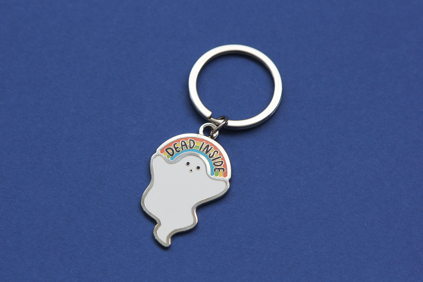 An enamel keychain showing a little white ghost holding a rainbow that says "Dead Inside" over a navy background.