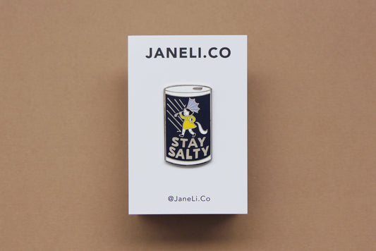 An enamel pin showing a grumpy girl on a salt can that says "Stay Salty" on a white JaneLi.Co backing card over a tan background.