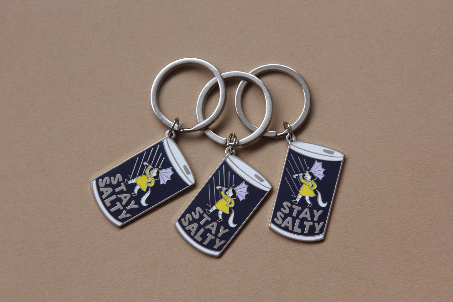 3 enamel keychains showing a grumpy girl on a salt can that says "Stay Salty" over a tan background.