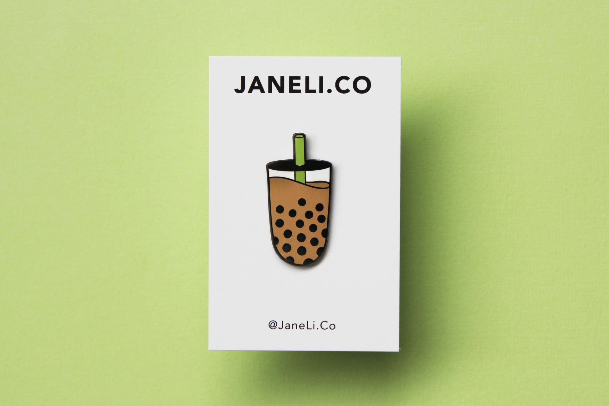 An enamel pin showing a cup of boba with a green straw on a white JaneLi.Co backing card over a green background.