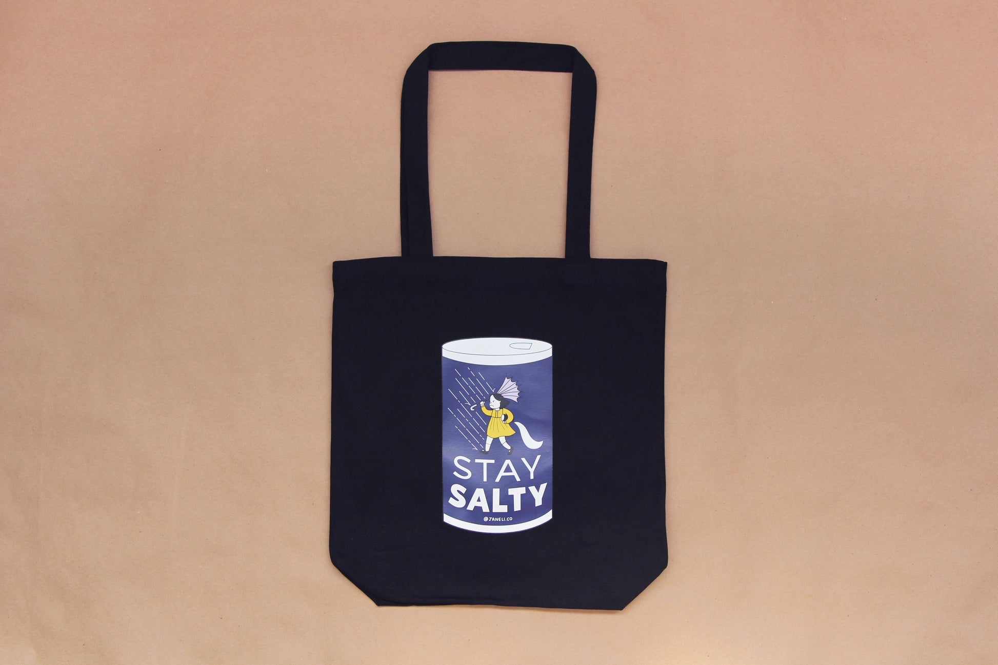 A photo of a black tote with a grumpy salt girl that says "Stay Salty" on it over a tan background.