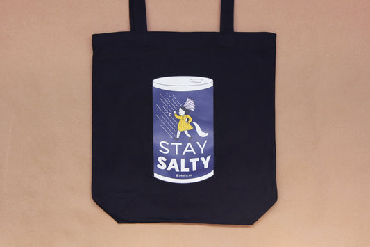 A photo of a black tote with a grumpy salt girl that says "Stay Salty" on it over a tan background.
