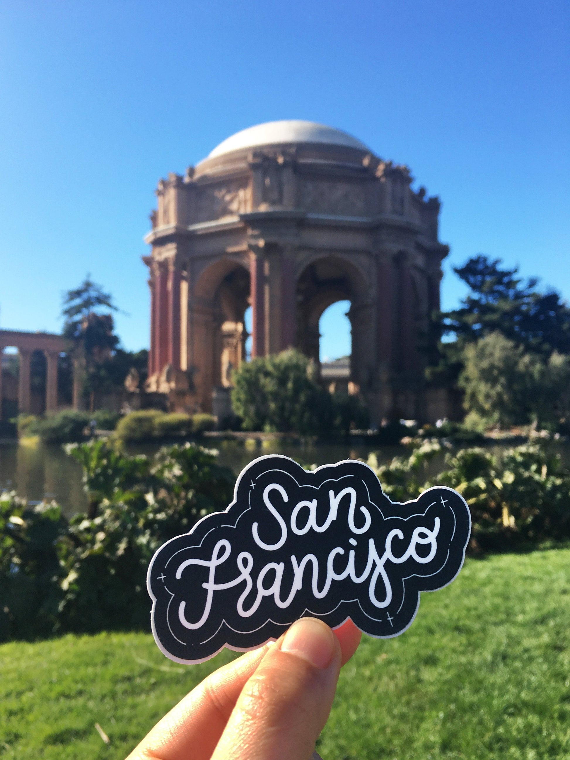 A hand holding a black and white JaneLi.Co sticker that says "San Francisco" in cursive lettering in front of the Palace of Fine Arts in San Francisco, California.