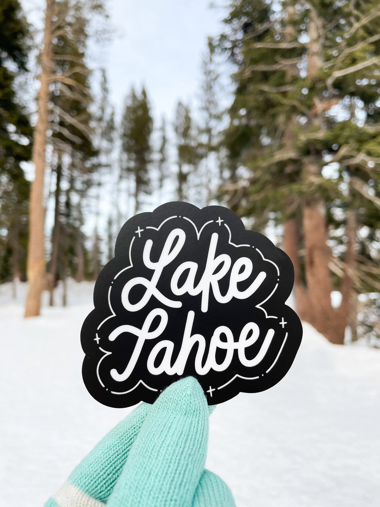 A gloved hand holding a black and white JaneLi.Co sticker that says "Lake Tahoe" in cursive lettering in front of the snowy outdoors.