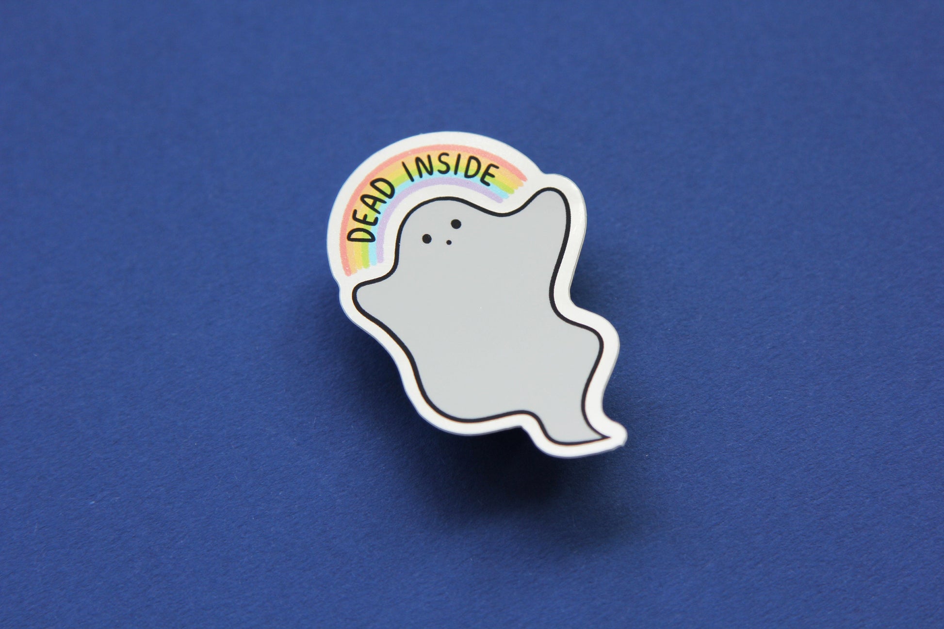 A JaneLi.Co sticker that shows a little white ghost holding up a rainbow that says "Dead Inside" over a navy blue background. The sticker has a metallic silver border.