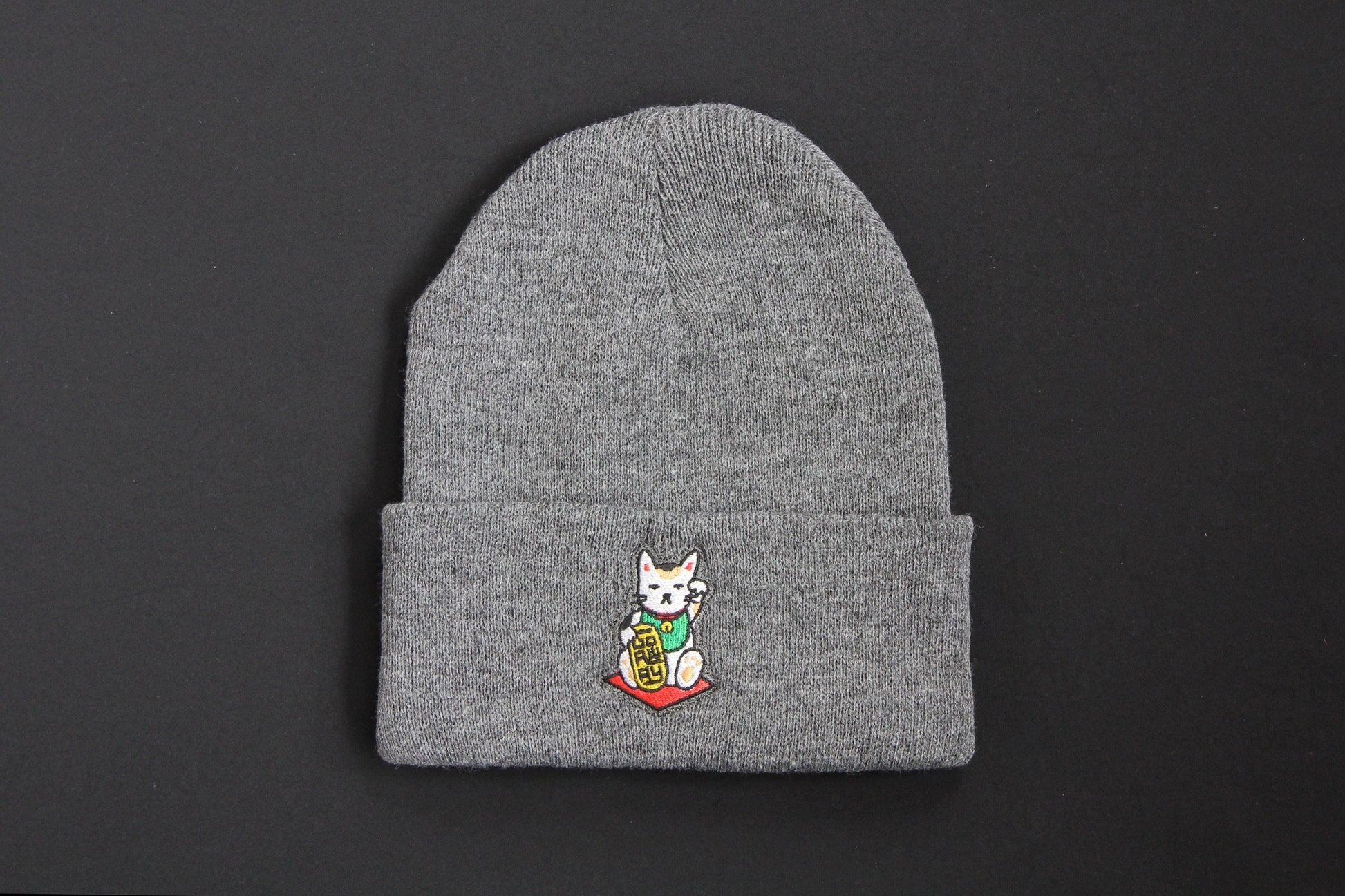 A cuffed charcoal grey beanie with an embroidered maneki neko cat holding a gold bar that says "Go Away" on a black background. 