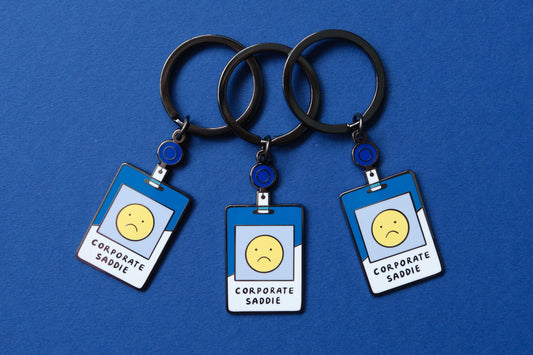 3 enamel keychains showing corporate badges that say "Corporate Saddie" with a sad face over a blue background.