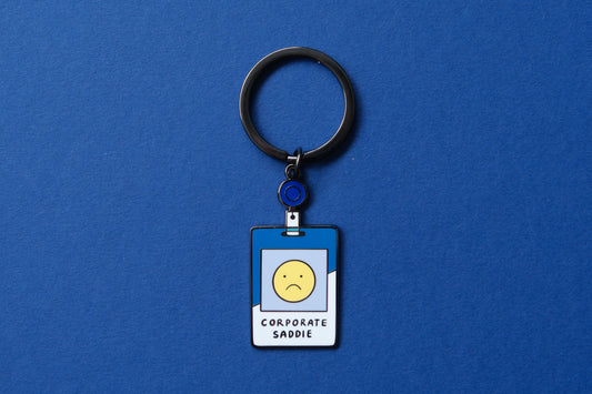 An enamel keychain showing a corporate badge that say "Corporate Saddie" with a sad face over a blue background.