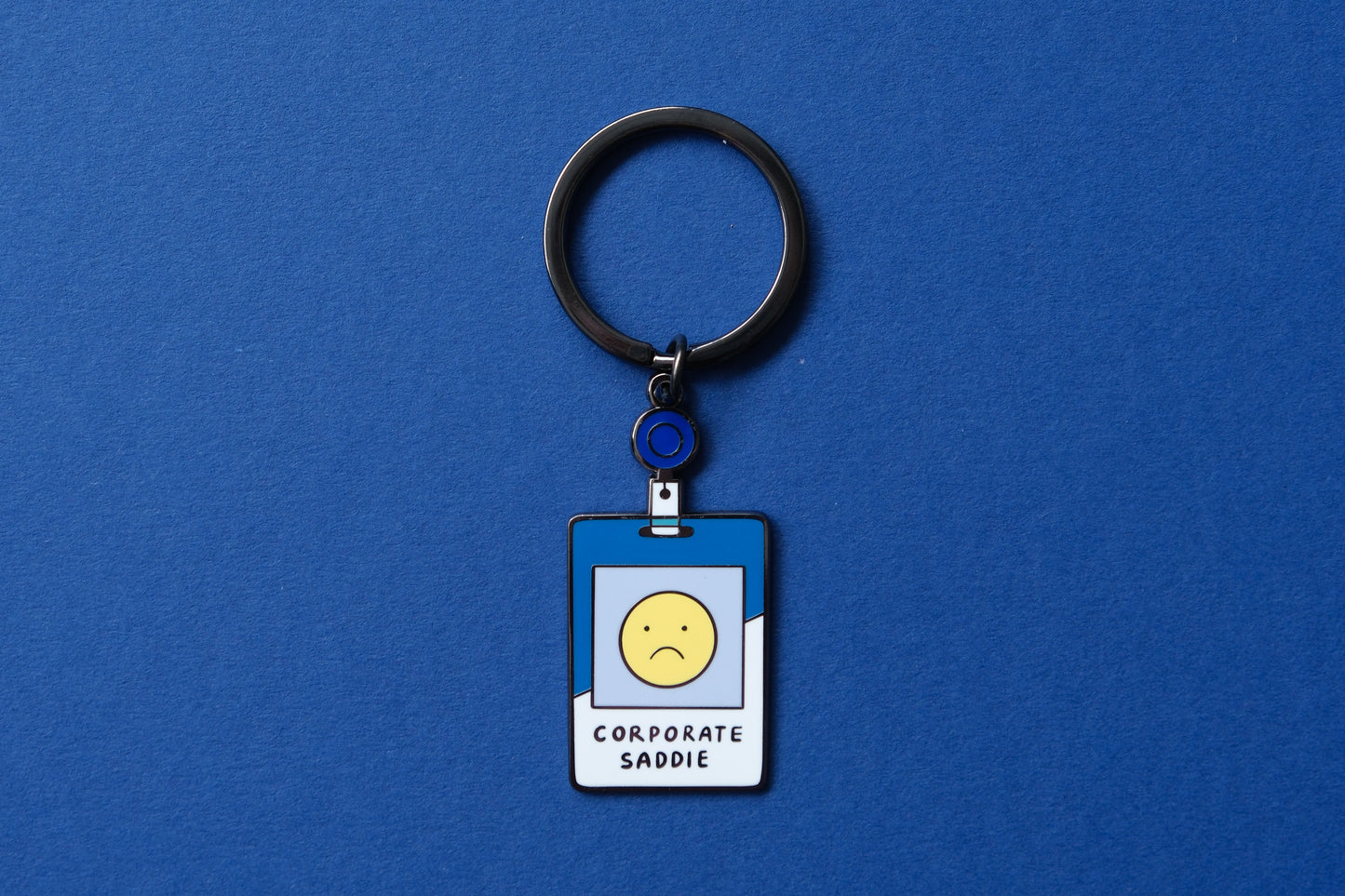 An enamel keychain showing a corporate badge that say "Corporate Saddie" with a sad face over a blue background.