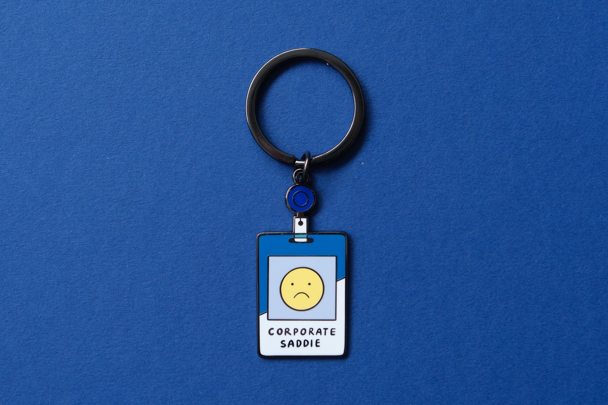 An enamel keychain showing a corporate badge that say "Corporate Saddie" with a sad face over a blue background.