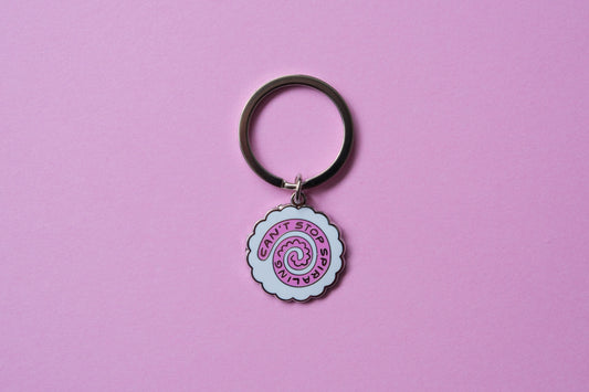 An enamel keychain showing a narutomaki fishcake that says "Can't Stop Spiraling" over a pink background.